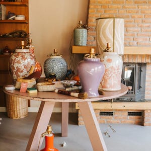 May include: A collection of decorative lamps and vases on a pink round table. The lamps have patterned shades and gold accents. Vases are in various colors and designs, including floral and geometric patterns. An orange lamp sits on the floor.