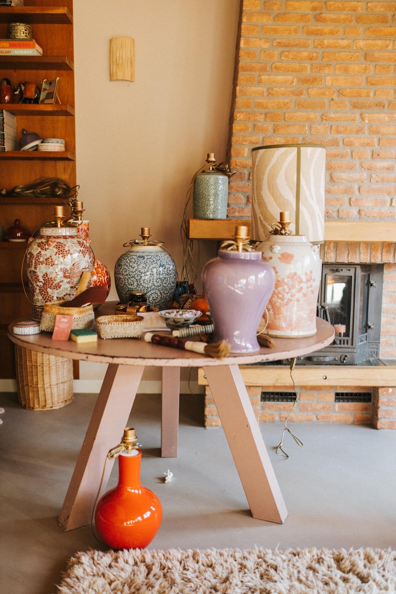May include: A collection of decorative lamps and vases on a pink round table. The lamps have patterned shades and gold accents. Vases are in various colors and designs, including floral and geometric patterns. An orange lamp sits on the floor.