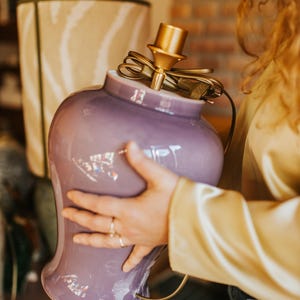 May include: A lavender-colored ceramic lamp base with a brass-colored lamp holder. The lamp base has a rounded shape with a flared base. A beige lampshade with a zebra stripe pattern is in the background.