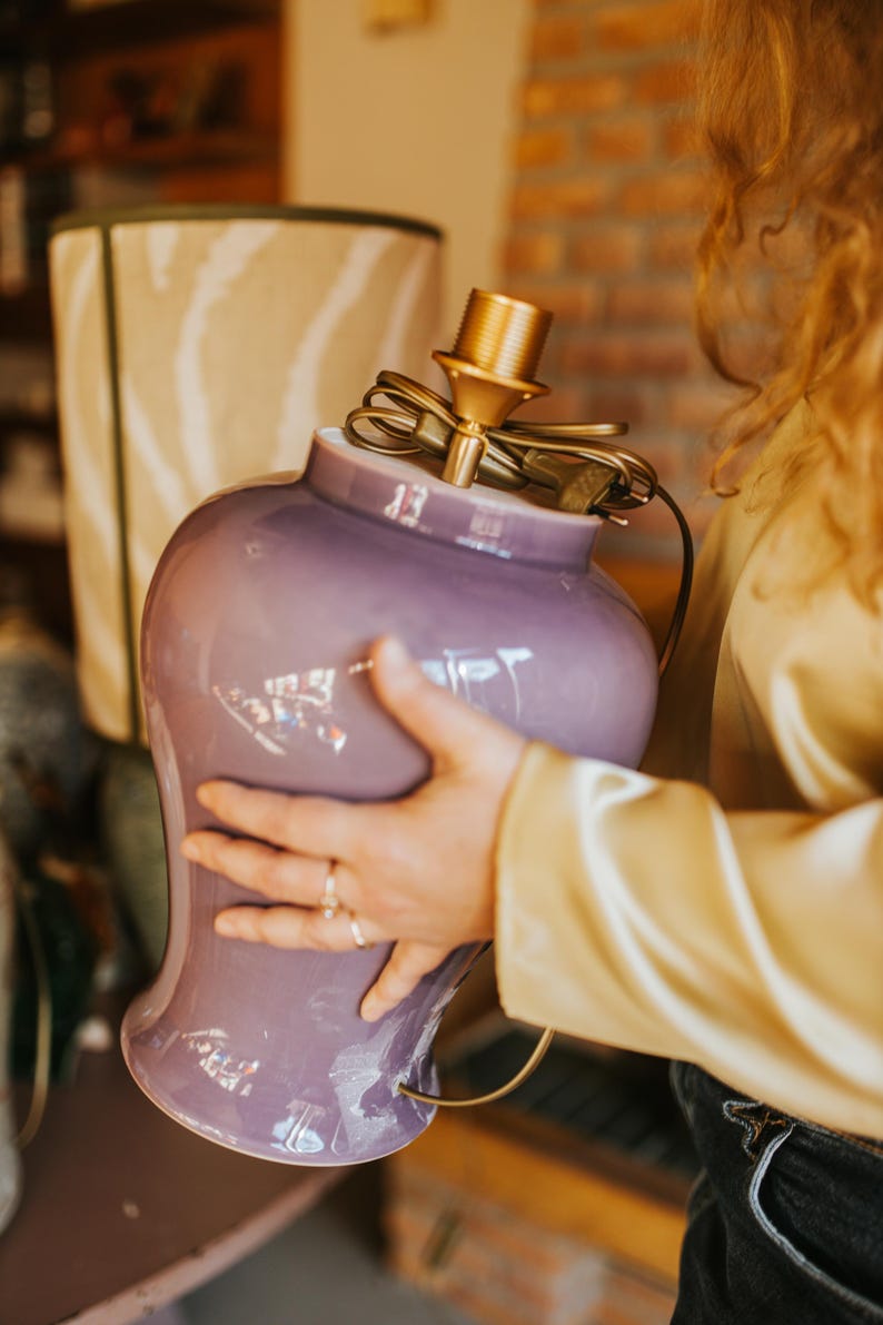 May include: A lavender-colored ceramic lamp base with a brass-colored lamp holder. The lamp base has a rounded shape with a flared base. A beige lampshade with a zebra stripe pattern is in the background.