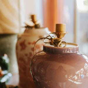 May include: A pair of decorative ceramic lamp bases. The front lamp is a glossy, mauve-colored jar-shaped base with a gold-colored lamp socket. The second lamp has a cream-colored base with a floral pattern. The lamps are on a wooden surface.