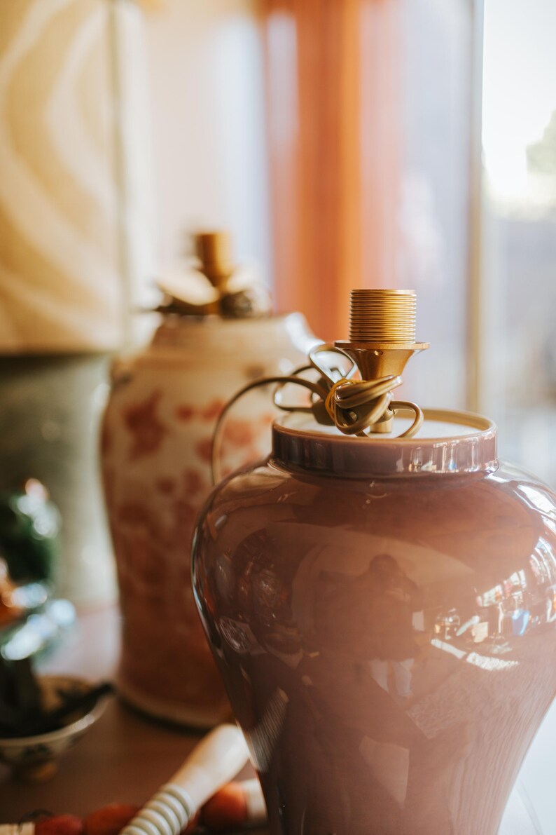 May include: A pair of decorative ceramic lamp bases. The front lamp is a glossy, mauve-colored jar-shaped base with a gold-colored lamp socket. The second lamp has a cream-colored base with a floral pattern. The lamps are on a wooden surface.