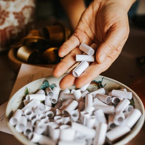 May include: A hand holds several small, white, cylindrical objects above a decorative bowl filled with similar items. The bowl has a floral and butterfly design. The objects appear to be small paper tubes.