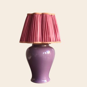 May include: A decorative table lamp with a lavender ceramic base and a pleated pink lampshade. The lampshade has a scalloped edge with a light brown trim. The lamp is designed for indoor use and provides ambient lighting.