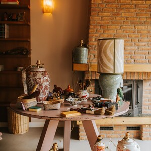 May include: A collection of decorative items displayed on a round, pink table. The items include ceramic lamps, vases, bowls, and baskets. A large lamp with a zebra-striped shade sits on the table. The background features a brick fireplace and wooden shelves.