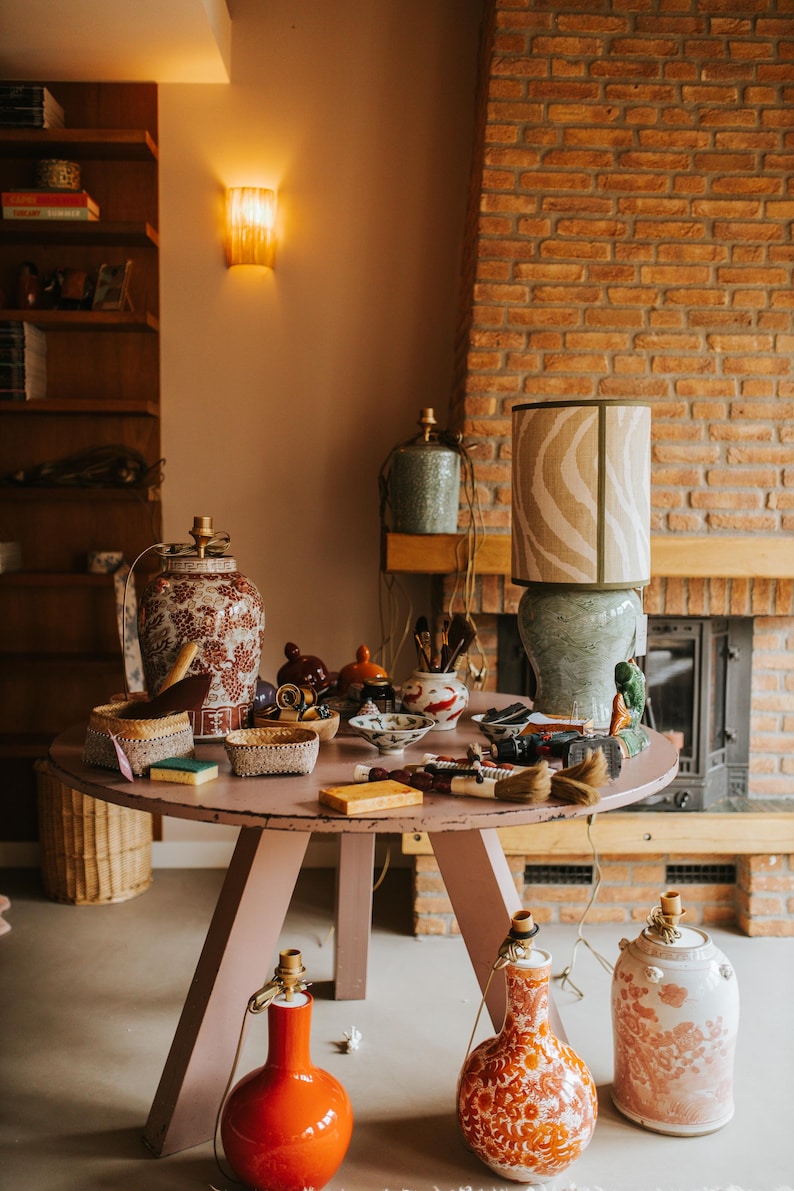 May include: A collection of decorative items displayed on a round, pink table. The items include ceramic lamps, vases, bowls, and baskets. A large lamp with a zebra-striped shade sits on the table. The background features a brick fireplace and wooden shelves.