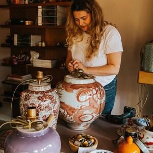 May include: A woman working on decorative ceramic lamps. The lamps are in various colors, including purple, red, and white, with intricate patterns. The lamps are on a pink table, with tools and other decorative items.