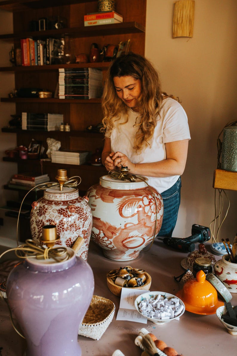 May include: A woman working on decorative ceramic lamps. The lamps are in various colors, including purple, red, and white, with intricate patterns. The lamps are on a pink table, with tools and other decorative items.