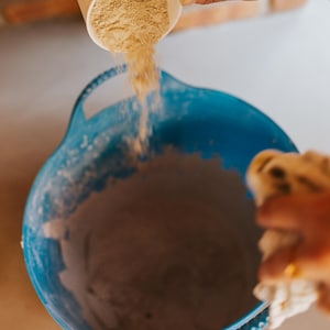 May include: A white mug with a brown illustration is pouring a light brown powder into a large blue bowl. The bowl is filled with a similar powder. The scene suggests baking or cooking.