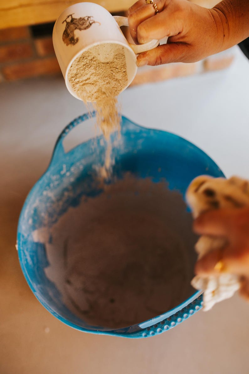 May include: A white mug with a brown illustration is pouring a light brown powder into a large blue bowl. The bowl is filled with a similar powder. The scene suggests baking or cooking.