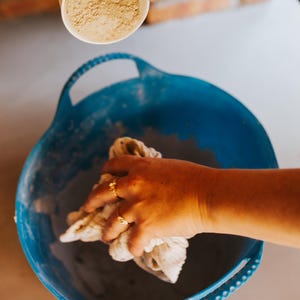 Peut inclure: Une tasse blanche avec une illustration de lapin verse une poudre beige dans un bassin bleu. Une main tient un chiffon blanc &agrave; l'int&eacute;rieur du bassin. L'image montre une activit&eacute; artisanale ou culinaire.