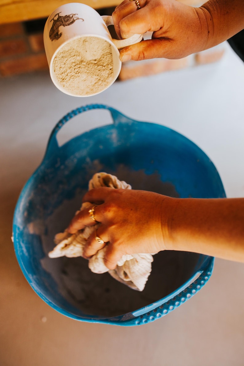 Peut inclure: Une tasse blanche avec une illustration de lapin verse une poudre beige dans un bassin bleu. Une main tient un chiffon blanc &agrave; l'int&eacute;rieur du bassin. L'image montre une activit&eacute; artisanale ou culinaire.