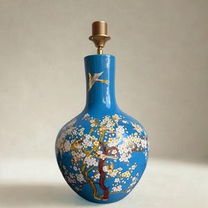 Könnte beinhalten: Eine blaue Keramik-Tischlampe mit einer goldfarbenen Lampenfassung. Die Lampe hat ein gemaltes Design eines blühenden Baumes mit weißen Blüten und Vögeln. Die Lampe ist etwa 30 cm hoch.