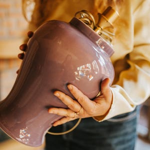 May include: A lavender-colored ceramic lamp base with a brass lamp holder. The lamp base has a rounded shape with a wide opening. The lamp is held by a person wearing a yellow shirt and blue jeans. The lamp is ready to be wired.