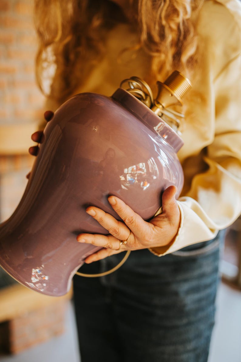 May include: A lavender-colored ceramic lamp base with a brass lamp holder. The lamp base has a rounded shape with a wide opening. The lamp is held by a person wearing a yellow shirt and blue jeans. The lamp is ready to be wired.