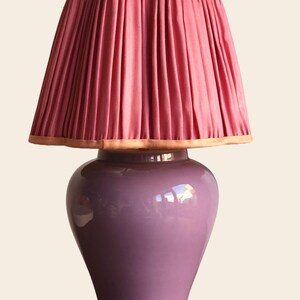 May include: A decorative table lamp with a pleated pink lampshade and a lavender-colored ceramic base. The lampshade has a scalloped edge with a light brown trim. The base has a glossy finish.