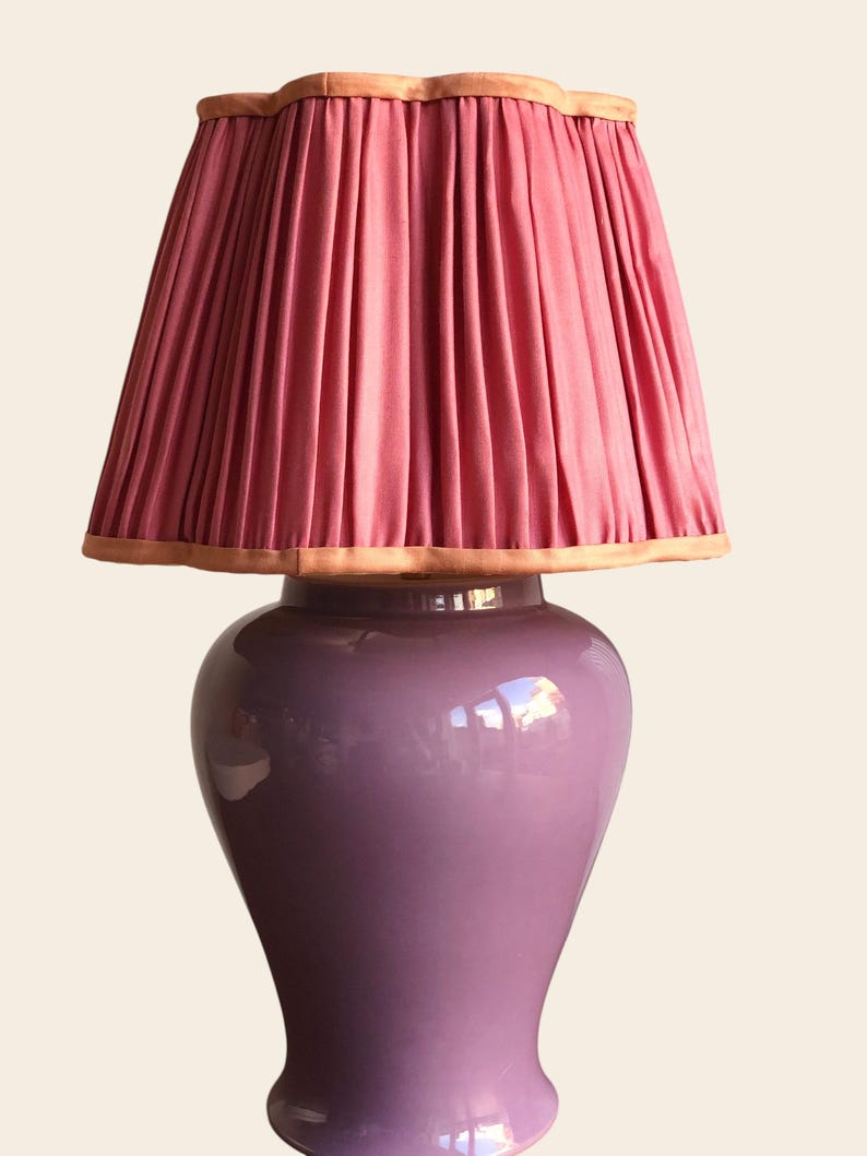 May include: A decorative table lamp with a pleated pink lampshade and a lavender-colored ceramic base. The lampshade has a scalloped edge with a light brown trim. The base has a glossy finish.
