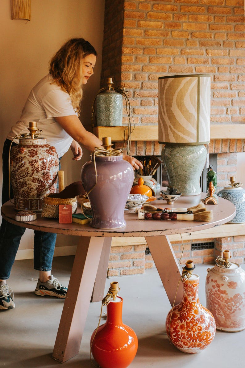 Peut inclure: Collection de lampes et de vases d&eacute;coratifs en c&eacute;ramique de diff&eacute;rentes couleurs et motifs, dont un grand vase orange, un vase lavande et des lampes avec des abat-jours &agrave; motifs. Les articles sont dispos&eacute;s sur une table ronde rose.