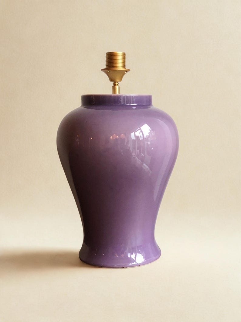 May include: A glossy, purple ceramic lamp base with a brass lamp holder. The lamp base has a rounded top and a flared base. The lamp is designed to hold a lampshade. The overall shape is reminiscent of a vase.
