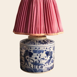 Puede incluir: Lámpara de mesa decorativa con una pantalla plisada rosa y un borde festoneado. La base es un jarrón de cerámica azul y blanco con un diseño de peces koi y nenúfares. Un elemento de decoración del hogar elegante.