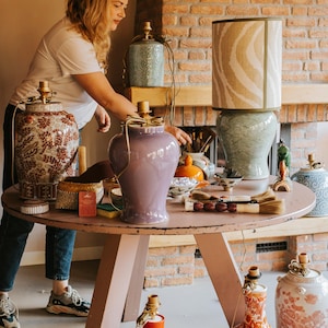 May include: A collection of decorative ceramic lamps and vases in various colors and patterns. The lamps have fabric shades, and the vases feature floral and geometric designs. The items are arranged on a round, pink table.