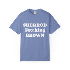 May include: A periwinkle blue t-shirt with the words "SHERROD F#@king BROWN" printed in white block letters. The shirt has short sleeves and a crew neck. The background is plain white.