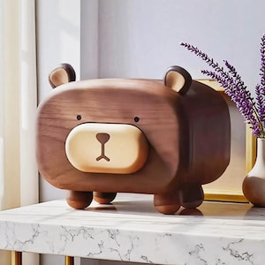 May include: A wooden bear-shaped storage box with a light brown face and dark brown body. The bear has small ears and a simple, friendly design. The box is sitting on a white marble surface.