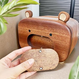 May include: A wooden bear-shaped box with a removable lid. The box is made of brown wood with two small, rounded ears on top and black dot eyes. The lid is being held open, revealing the interior.