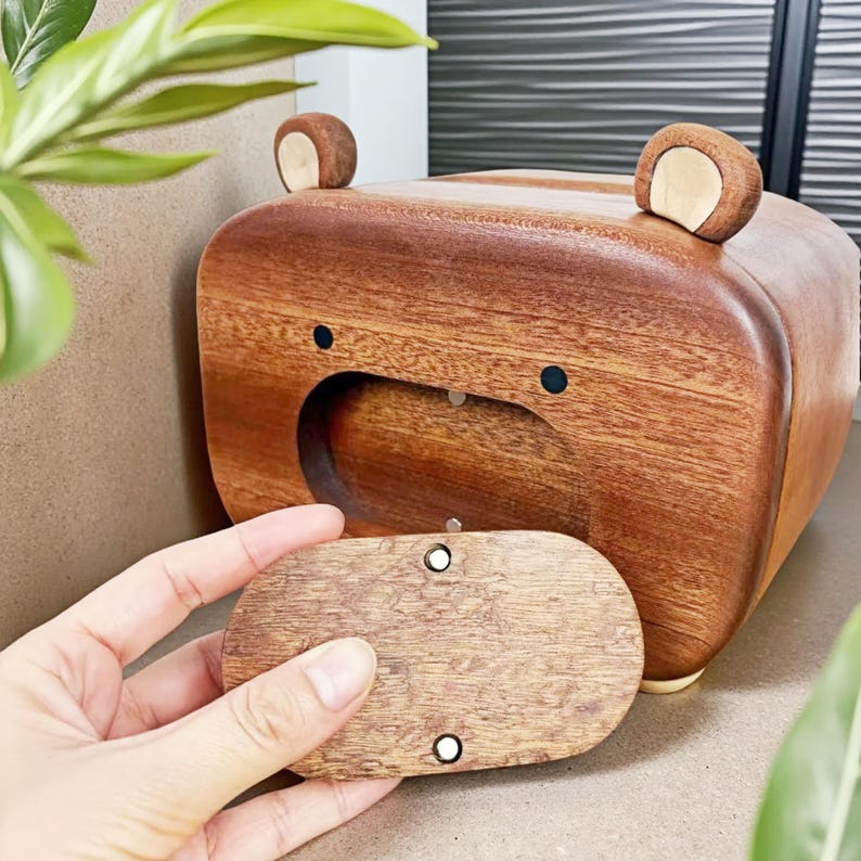 May include: A wooden bear-shaped box with a removable lid. The box is made of brown wood with two small, rounded ears on top and black dot eyes. The lid is being held open, revealing the interior.