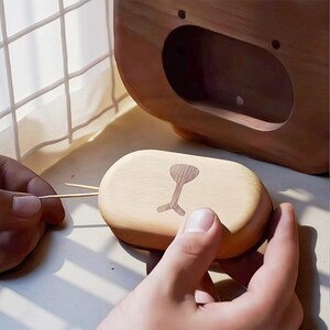 May include: A light-colored wooden toothpick holder with a bear face design. The holder is oval-shaped with a carved bear face on the lid. The image shows the lid being opened, revealing the toothpicks inside. The holder is made of light-colored wood.