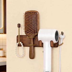 May include: A wall-mounted dark wood organizer rack featuring a brass hook holding a white beaded bracelet, a central brass ring support for a wooden hairbrush with bristles, and two wooden pegs supporting a white hair dryer with a power cord.