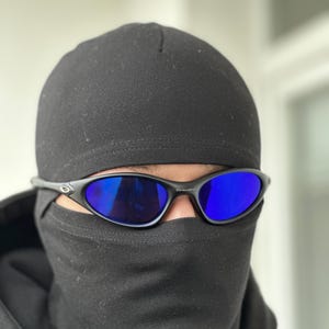 K&ouml;nnte beinhalten: Schwarze Sonnenbrille mit blauen Gl&auml;sern wird mit einer schwarzen M&uuml;tze und Gesichtsbedeckung getragen. Die Sonnenbrille hat ein schlankes, sportliches Design. Die Gl&auml;ser reflektieren eine leuchtend blaue Farbe, die mit der dunklen Kopfbedeckung kontrastiert.