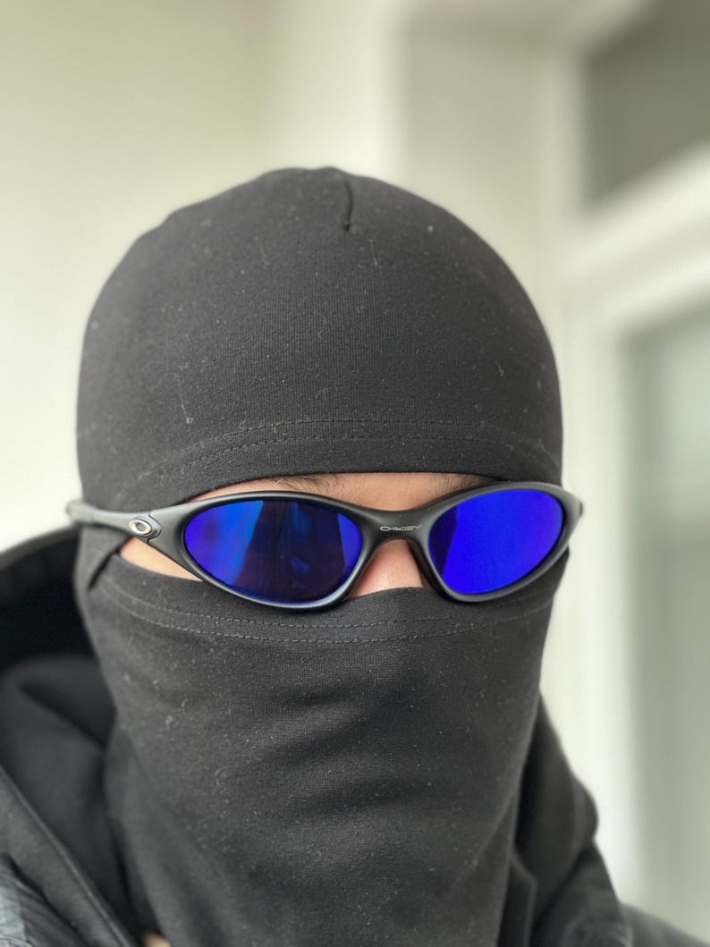 K&ouml;nnte beinhalten: Schwarze Sonnenbrille mit blauen Gl&auml;sern wird mit einer schwarzen M&uuml;tze und Gesichtsbedeckung getragen. Die Sonnenbrille hat ein schlankes, sportliches Design. Die Gl&auml;ser reflektieren eine leuchtend blaue Farbe, die mit der dunklen Kopfbedeckung kontrastiert.
