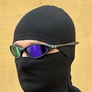 May include: Black sunglasses with a dark frame and mirrored lenses are worn with a black balaclava. The balaclava covers the head and neck, with only the eyes visible. The background is a solid, light brown color.