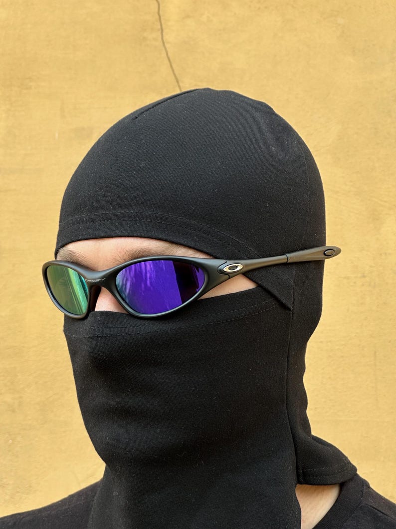 May include: Black sunglasses with a dark frame and mirrored lenses are worn with a black balaclava. The balaclava covers the head and neck, with only the eyes visible. The background is a solid, light brown color.