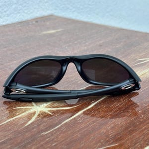 May include: Black sunglasses with dark lenses. The sunglasses have a sleek, modern design with a curved frame and a bridge. The arms of the sunglasses are folded. The sunglasses are on a wooden surface.