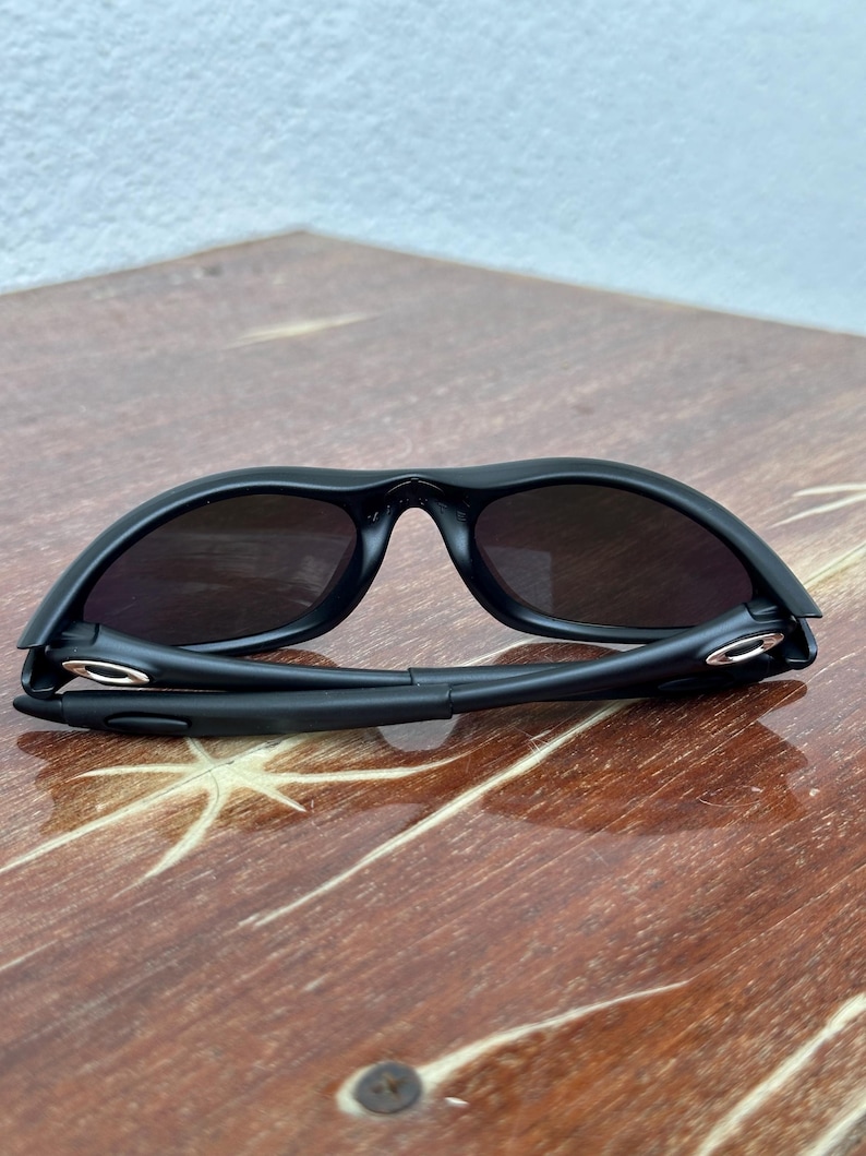 May include: Black sunglasses with dark lenses. The sunglasses have a sleek, modern design with a curved frame and a bridge. The arms of the sunglasses are folded. The sunglasses are on a wooden surface.