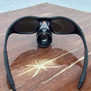 May include: Black sunglasses with dark lenses. The sunglasses have a sleek, sporty design with curved frames and a bridge. The lenses are oval-shaped and appear to be tinted. The sunglasses are resting on a wooden surface.