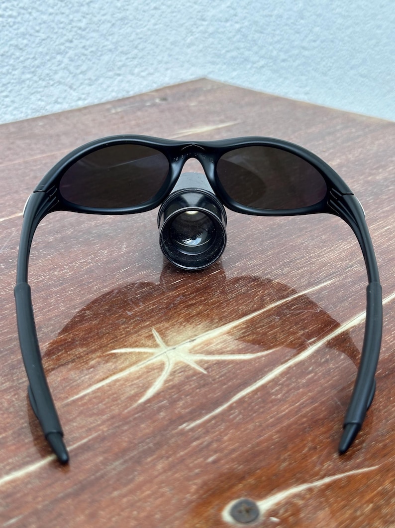May include: Black sunglasses with dark lenses. The sunglasses have a sleek, sporty design with curved frames and a bridge. The lenses are oval-shaped and appear to be tinted. The sunglasses are resting on a wooden surface.