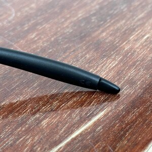 May include: A close-up of a black stylus pen with a rounded body and a pointed tip. The pen is lying on a wooden surface with a warm, brown tone. The pen's tip is angled towards the lower right corner.