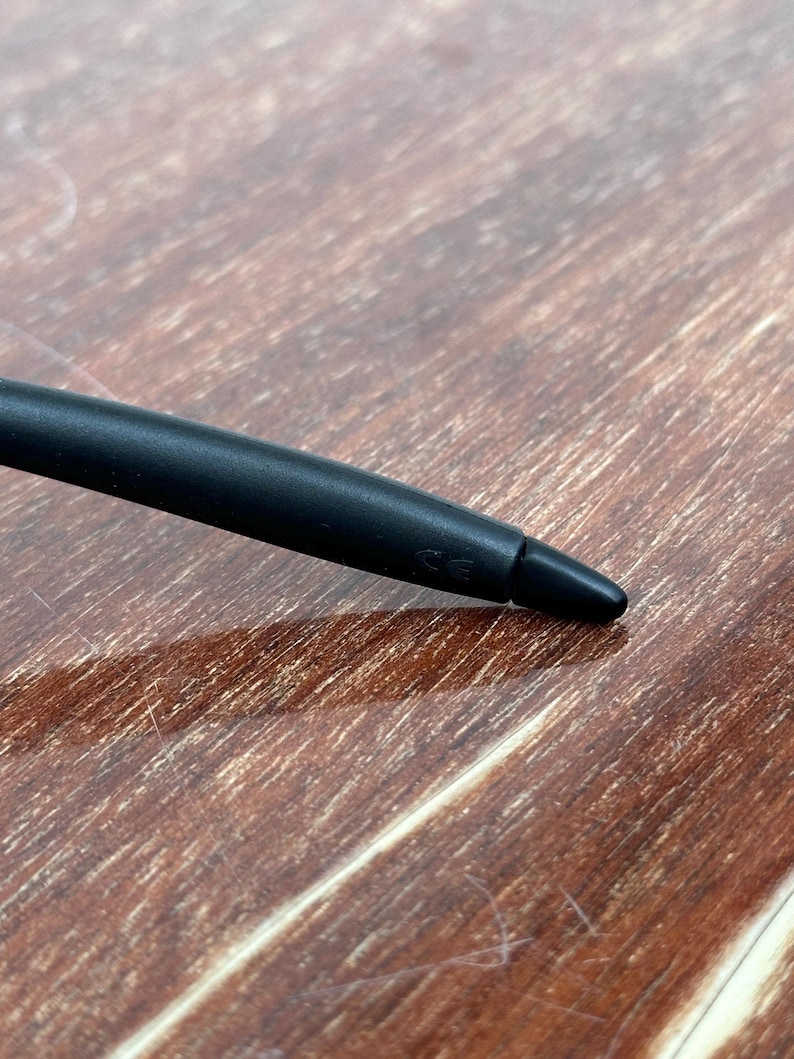 May include: A close-up of a black stylus pen with a rounded body and a pointed tip. The pen is lying on a wooden surface with a warm, brown tone. The pen's tip is angled towards the lower right corner.