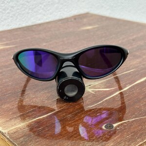 May include: Black Oakley sunglasses with purple-tinted lenses. The sunglasses are resting on a black cylindrical object. The lenses reflect a blue and purple hue. The frame is black and curved.