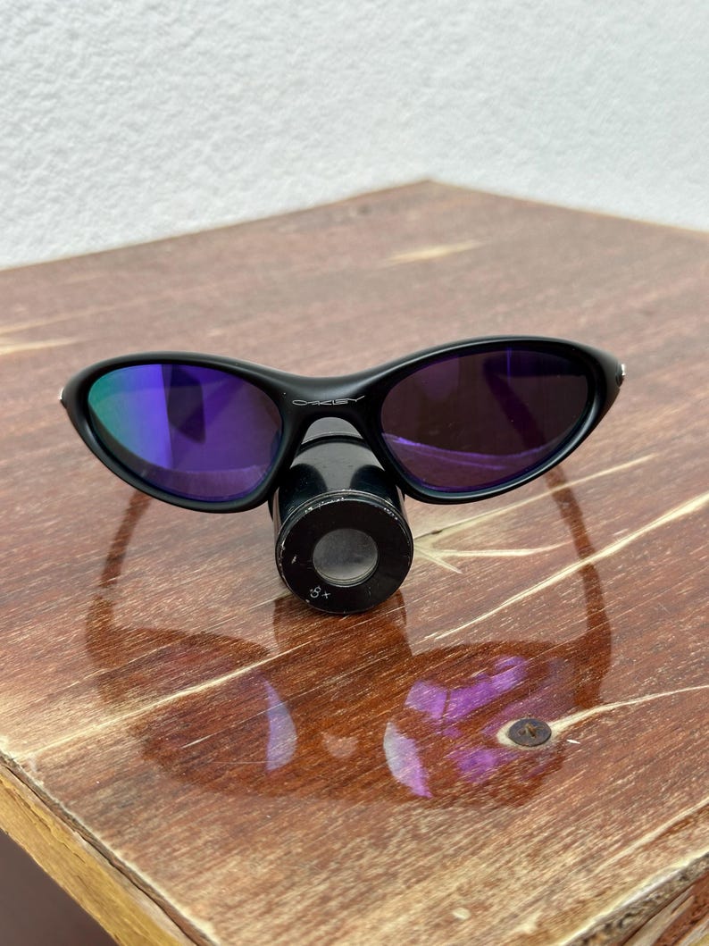 May include: Black Oakley sunglasses with purple-tinted lenses. The sunglasses are resting on a black cylindrical object. The lenses reflect a blue and purple hue. The frame is black and curved.