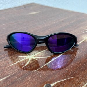 May include: Black Oakley sunglasses with purple mirrored lenses rest on a wooden surface. The sunglasses have a sleek, curved frame design. The lenses reflect the surrounding environment, creating a vibrant, colorful effect. The Oakley logo is visible on the frame.