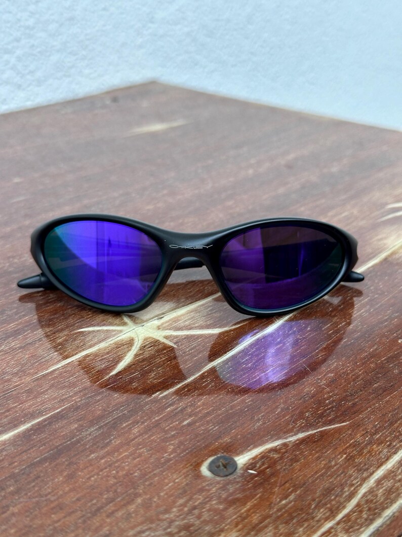 May include: Black Oakley sunglasses with purple mirrored lenses rest on a wooden surface. The sunglasses have a sleek, curved frame design. The lenses reflect the surrounding environment, creating a vibrant, colorful effect. The Oakley logo is visible on the frame.
