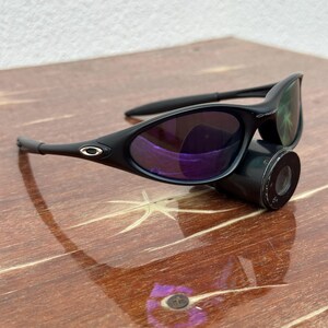 May include: Black sunglasses with purple-tinted lenses. The sunglasses have a sleek, curved design with a matte finish. The frame features a small silver logo on the temples. The lenses reflect a purple hue, and the arms are thin and curved.