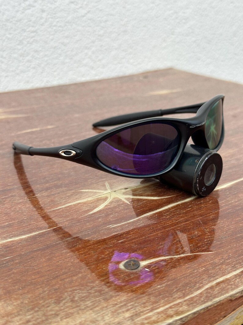 May include: Black sunglasses with purple-tinted lenses. The sunglasses have a sleek, curved design with a matte finish. The frame features a small silver logo on the temples. The lenses reflect a purple hue, and the arms are thin and curved.