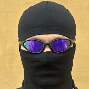 May include: Black sunglasses with purple mirrored lenses are worn over a black balaclava and a black cap. The balaclava covers the lower face and neck. The background is a solid, light brown color.