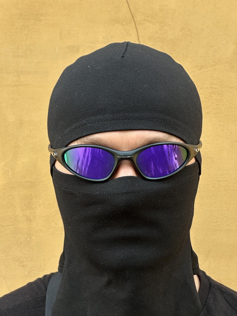 May include: Black sunglasses with purple mirrored lenses are worn over a black balaclava and a black cap. The balaclava covers the lower face and neck. The background is a solid, light brown color.