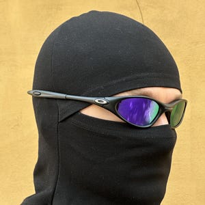 May include: Black sunglasses with purple-tinted lenses are worn over a black balaclava. The sunglasses have a thin, dark frame. The background is a solid, light brown color.